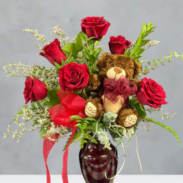 Red roses arranged in a vase with a teddy bear and ribbon accents