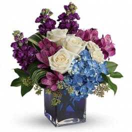 Bouquet of white roses, blue hydrangeas, and purple flowers in a blue glass vase