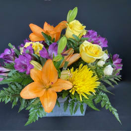 Orange lilies, yellow roses, and purple alstroemeria in a rectangular vase