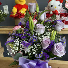 Lavender roses and lilies arranged with purple filler flowers and a satin ribbon bow.