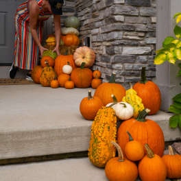 A woman arranges a stack of pumpkins and gourds on a front porch.