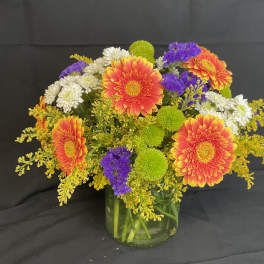 Mixed bouquet of orange gerbera daisies, white mums, and purple filler in a glass vase