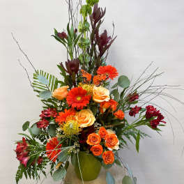 Tall floral arrangement with orange roses, gerbera daisies, and burgundy blooms in a green vase