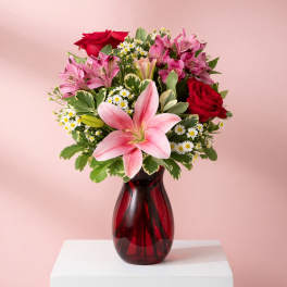 Bouquet of pink lilies, red roses, and small white daisies in a red glass vase