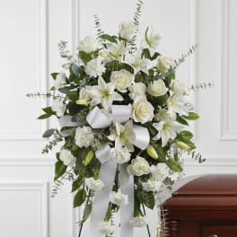 Standing spray of white roses, lilies, and carnations with a white ribbon on an easel beside a wooden casket.