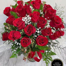 Tall bouquet of red roses with white filler flowers in a clear glass vase with a red ribbon