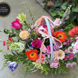 Colorful basket arrangement of pink roses, orange gerbera daisies, and mixed blooms with pastel ribbon bows