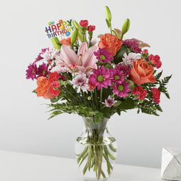 Mixed bouquet of pink lilies, roses, and daisies in a glass vase with a birthday pick
