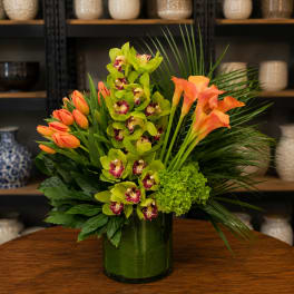 Tropical arrangement with orange tulips, green orchids, and calla lilies in a glass vase