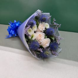 Bouquet of pale roses, white blooms, and blue thistle wrapped in lavender paper