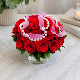 Red roses arranged in a clear glass vase with pink ribbon and pearl strands