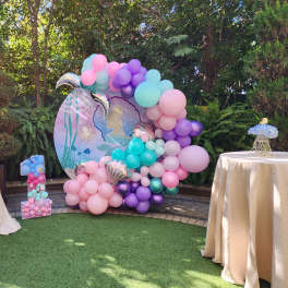 Pastel balloon display with a mermaid backdrop and two draped tables outdoors.