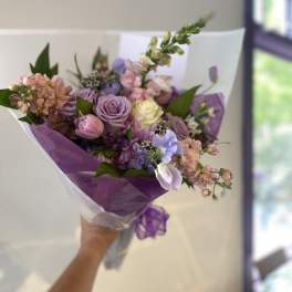 Handheld bouquet of lavender, pink, and cream flowers wrapped in purple paper