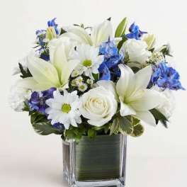 White lilies, roses, and daisies with blue flowers in a square glass vase