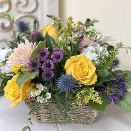 Mixed flower arrangement in a woven basket with yellow roses and purple blooms