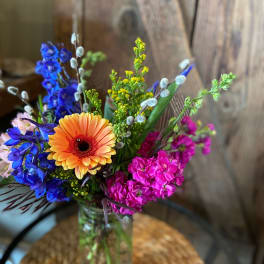 Colorful arrangement with orange gerbera, blue delphinium, and pink stock in a clear glass vase