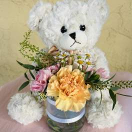 White teddy bear holding a small flower arrangement in a glass vase