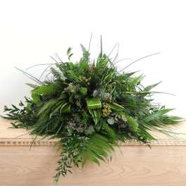 Large low arrangement of assorted green foliage on a wooden casket