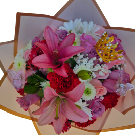 Wrapped bouquet of pink lilies, roses, red carnations and white daisies with a small gold crown pick.