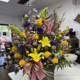 Tall mixed bouquet in a woven basket with pink ribbon and yellow lilies