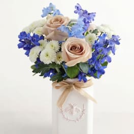 Bouquet of blush roses, blue flowers, and white mums in a white vase with a ribbon
