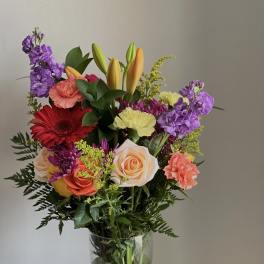 Mixed bouquet in a clear glass vase with roses, lilies, carnations, and purple blooms