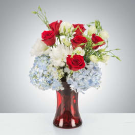 Red roses and white flowers arranged with blue hydrangeas in a red glass vase.