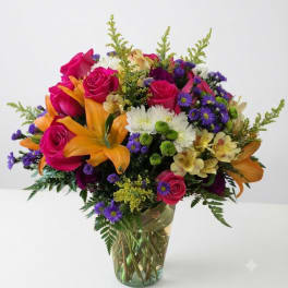 Mixed bouquet of pink roses, orange lilies, and purple daisies in a glass vase