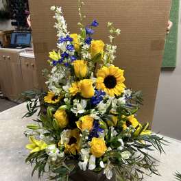 Tall floral arrangement with yellow roses, sunflowers, and white blooms in a dark box.