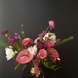 Asymmetrical arrangement of pink and purple flowers in a gold pedestal vase against a dark background