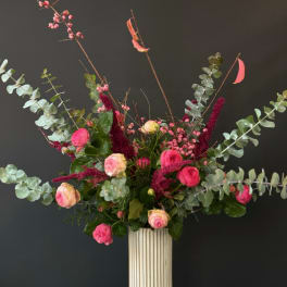 Tall arrangement of pink roses, berries, and burgundy accents in a white ribbed vase with sweeping eucalyptus.