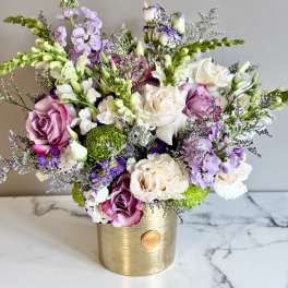 Lavender, white, and pink mixed blooms arranged in a gold cylinder vase on a marble surface.