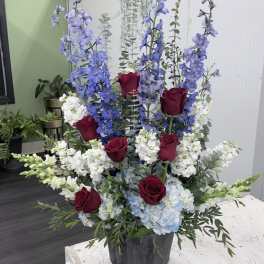 Tall floral arrangement with red roses, blue delphinium, and white blooms in a black container