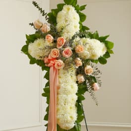 Standing floral cross with white blooms, peach roses, and a pink ribbon