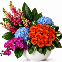 Mixed bouquet in a white bowl vase with orange roses, blue hydrangeas, pink snapdragons, and purple orchids