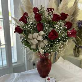 Tall arrangement of red roses with white filler flowers in a red glass vase with a gold bow