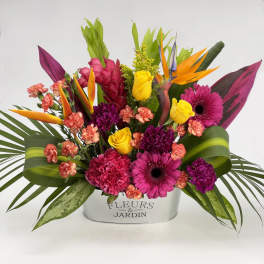 Bright tropical arrangement with pink gerbera daisies, yellow roses, and orange blooms in a metal container.