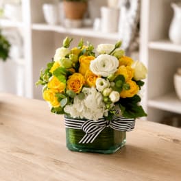 Yellow and white flower arrangement in a square glass vase with a striped ribbon