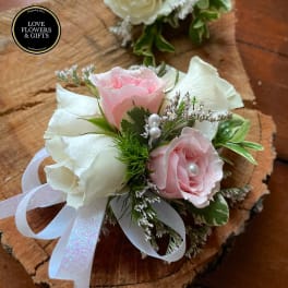 Pink and white rose wrist corsage with ribbon and pearl accents on a wood slice