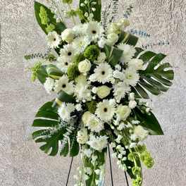Tall standing spray of white roses, gerbera daisies, and mums with large tropical leaves on an easel