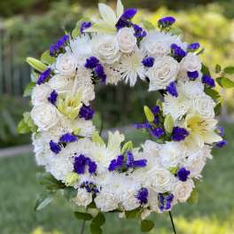 Open standing wreath of white roses, mums, and lilies with bright purple accents on a metal stand.