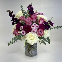 Mixed bouquet of white roses, pink roses, and purple flowers in a gray textured glass vase