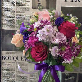 Mixed bouquet of pink, purple, and peach flowers in a glass vase with a purple ribbon
