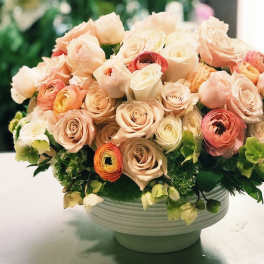 Round bouquet of blush and cream roses with green accents in a white bowl vase