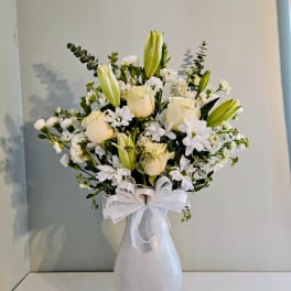 White roses and lilies arranged in a white vase with a ribbon bow