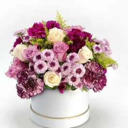 Round arrangement of cream and lavender roses, purple carnations, and mums in a white hatbox container