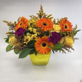 Bright arrangement of orange gerbera daisies, yellow roses, and purple blooms in a lime-green square vase