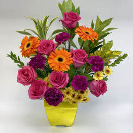 Bright arrangement of pink roses, orange gerbera daisies and purple carnations in a yellow square vase