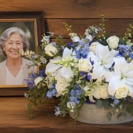 White lilies and roses in a low basket beside a memorial urn and framed portrait