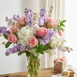 Pink roses and lavender flowers in a clear glass vase with gift boxes nearby